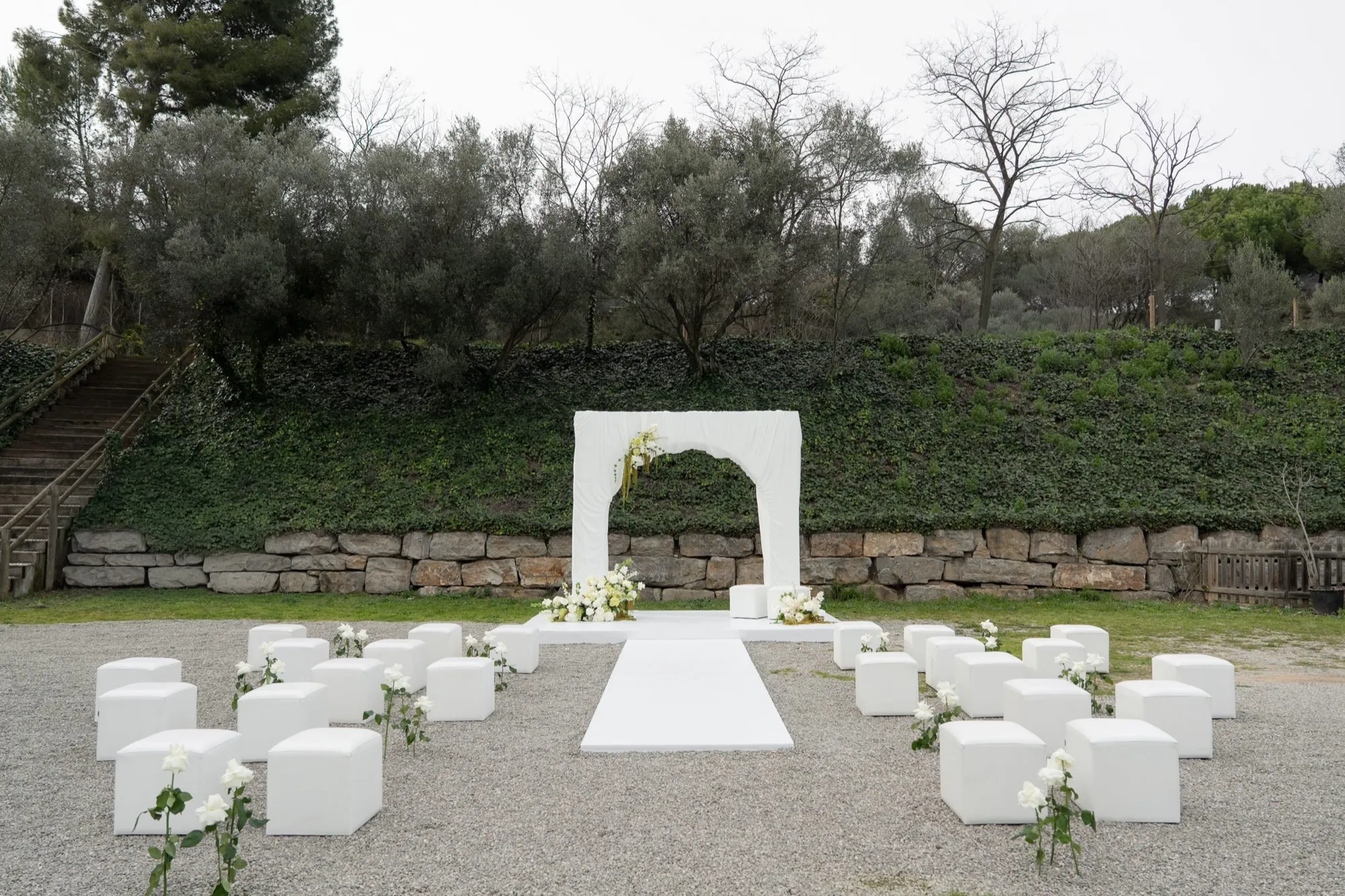 Ceremonia en el jardín con arco blanco y asientos entre olivos