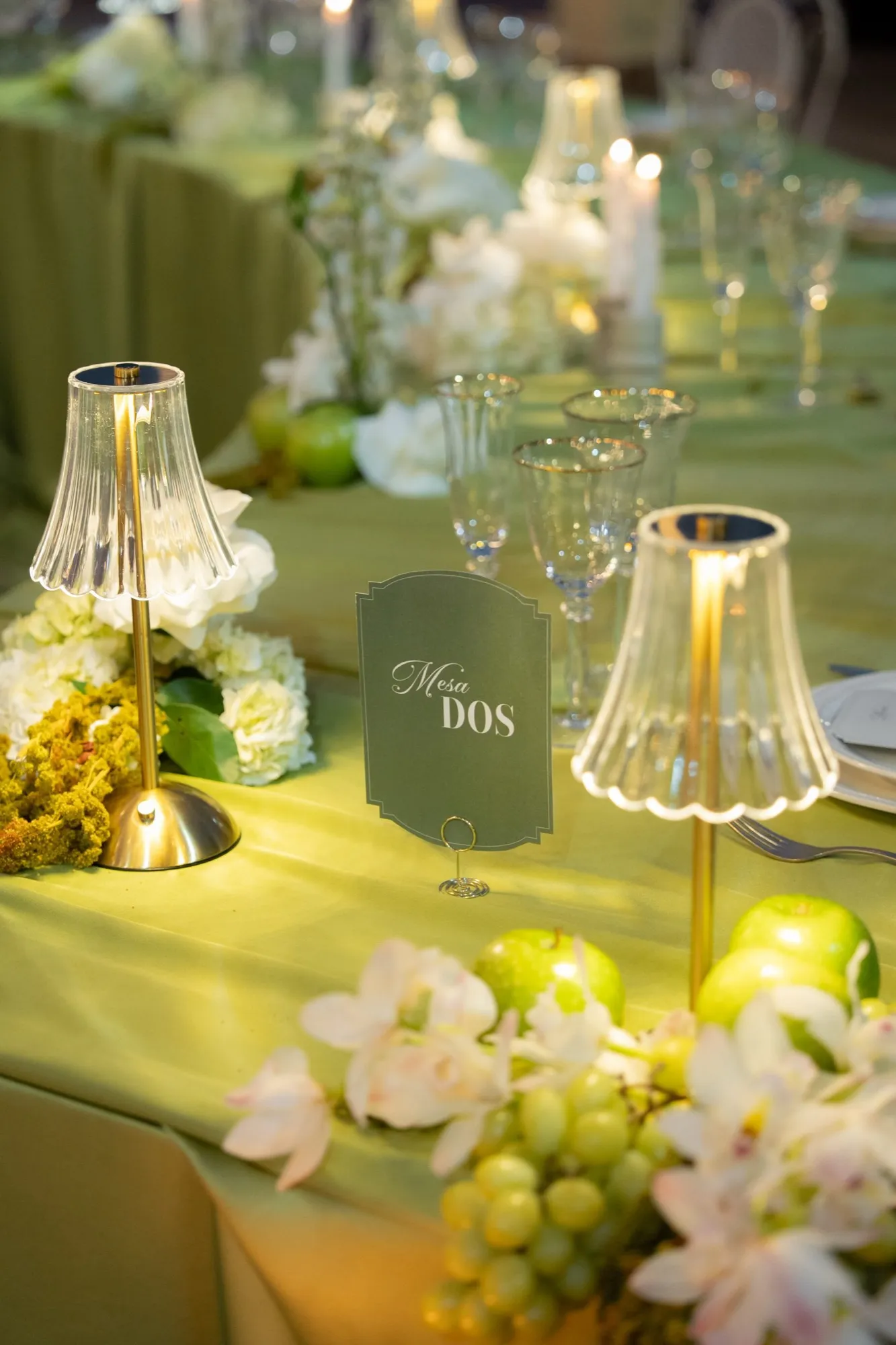 Mesa de boda con mantel verde oliva, flores blancas y lámparas