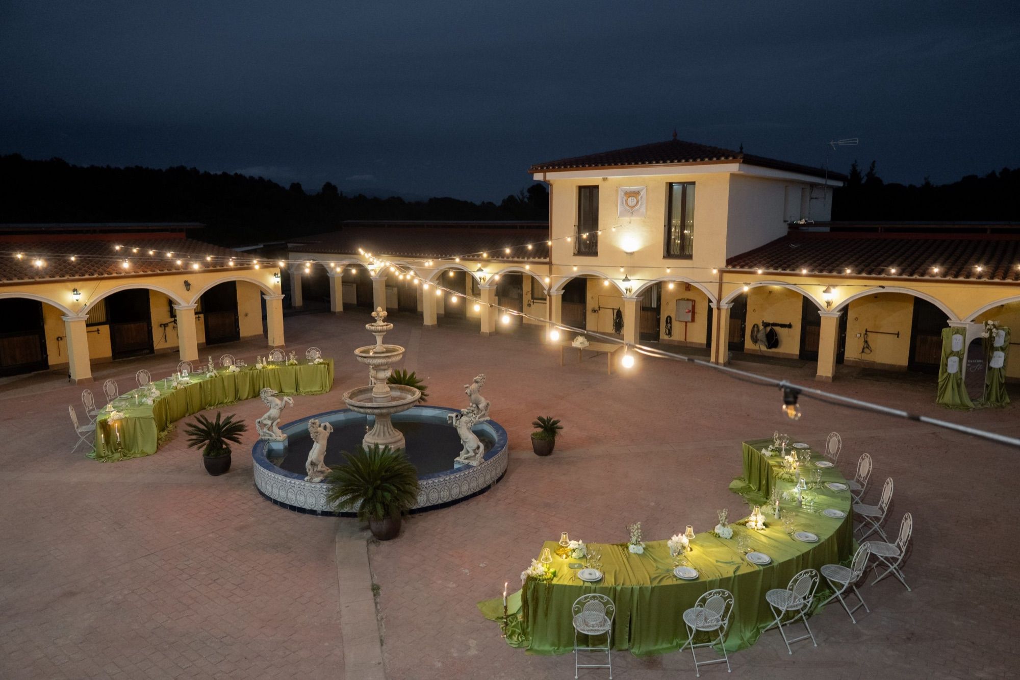 Patio de la finca Binomio Ecuestre de noche, con la fuente iluminada, guirnaldas de luces y mesas de boda en verde oliva